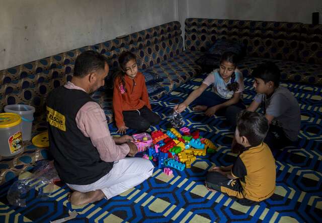 Eine Gruppe von Menschen sitzt auf einem blauen gemusterten Boden; ein Erwachsener in einer Weste interagiert mit Kindern, die mit buntem Spielzeug spielen, in einem einfachen, gemütlichen Raum.